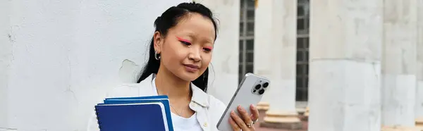 A pretty young Asian student stands near her university, engaged with her phone and holding books. — Stock Photo