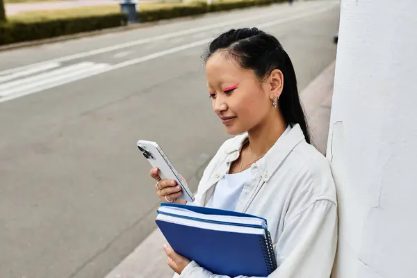 A stylish young student stands on campus, holding her notebooks and smiling at her phone. — Stock Photo
