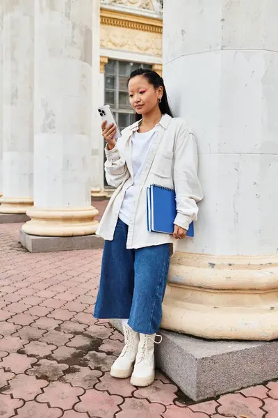 A young Asian woman in a fashionable outfit is using her smartphone while standing by university columns. — Stock Photo