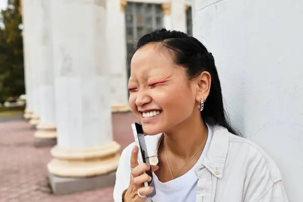 A cheerful young Asian student in a stylish outfit laughs while leaning against a university column. — Stock Photo