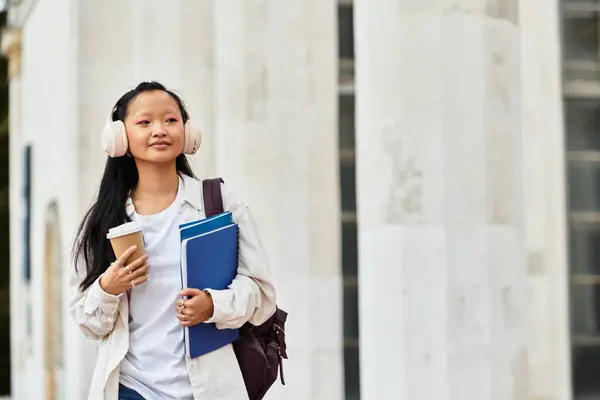 A young Asian student in a fashionable outfit walks with coffee and textbooks, embracing campus life vividly. — Stock Photo