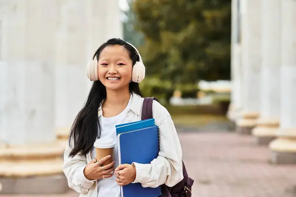 A stylish young student girl smiles as she walks through the university campus with coffee and books. — Stock Photo