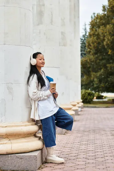 A young Asian student stands near university columns, sipping coffee and embracing campus life with style. — Stock Photo
