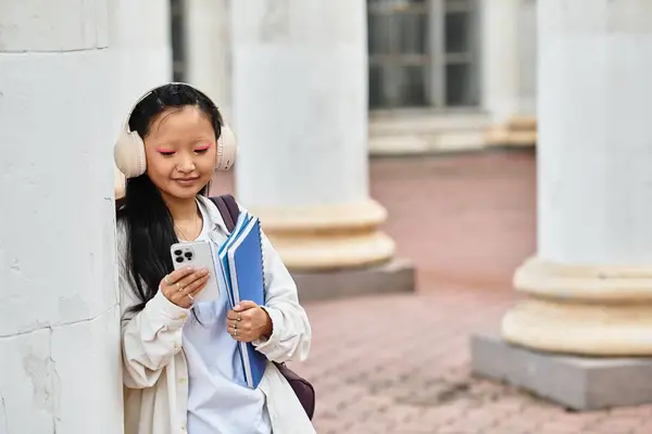 A stylish young Asian student enjoys her university campus, happily checking her phone with books in hand. — Stock Photo