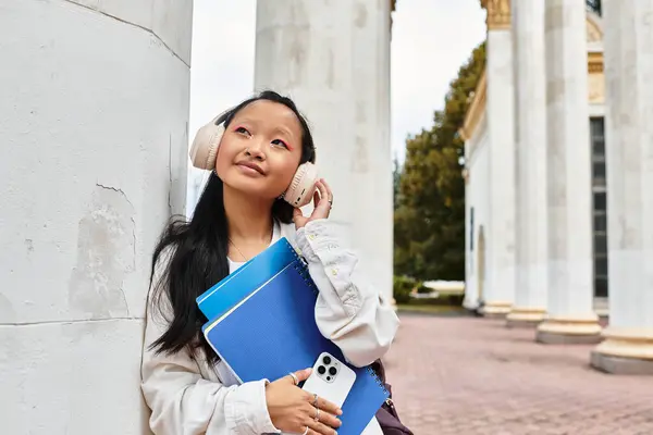A fashionable young woman relaxes with headphones, holding notebooks and a phone in a university setting. — Stock Photo