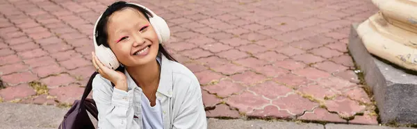 A stylish young girl with headphones smiles while lounging on the campus path, embracing student life. — Stock Photo