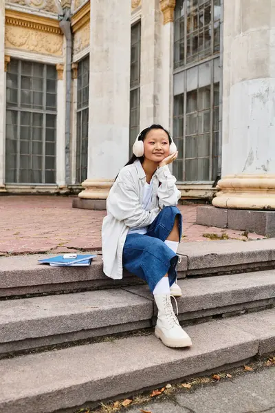 A young Asian student dressed in a stylish outfit relaxes on campus steps, embracing autumn vibes with warmth. — Stock Photo