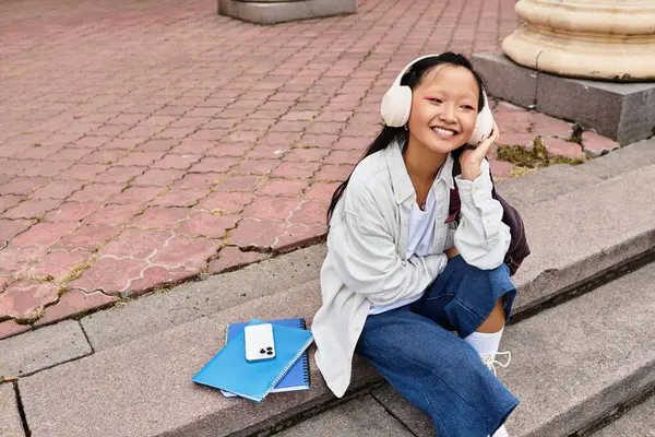 A cheerful young student sits on steps with notebooks and headphones, embracing youth culture on campus. — Stock Photo