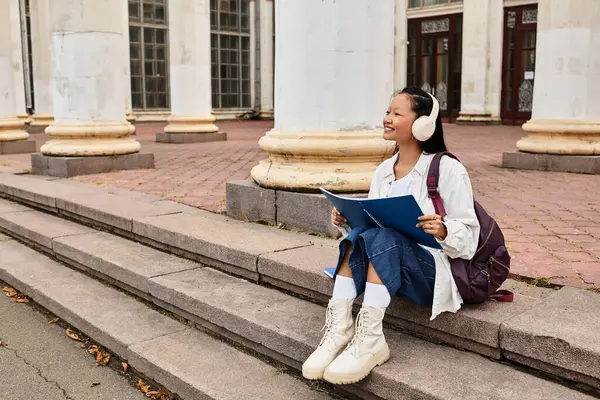 Una giovane ragazza asiatica in un abito elegante studia felicemente sui gradini del campus, immergendosi nella vita universitaria e nella cultura.. — Foto stock
