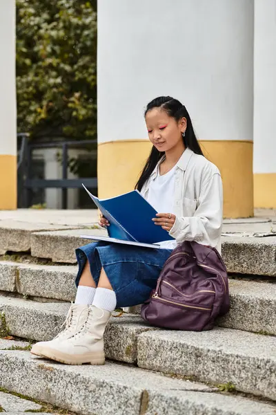 A young Asian student sits on steps, reading intently while dressed stylishly with a backpack beside her. — Stock Photo