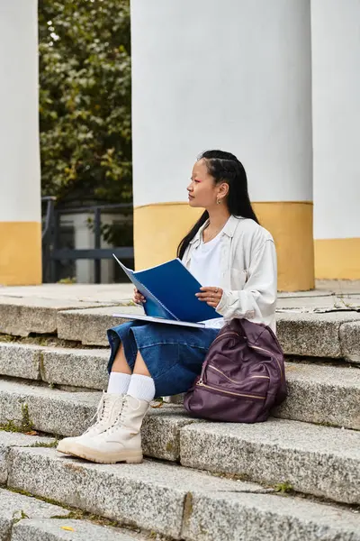 A young Asian student sits on university steps, studying her notes, showcasing modern youth fashion and culture. — Stock Photo