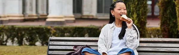 A cheerful young Asian student in a fashionable outfit eating sandwich while relaxing on a bench. — Stock Photo