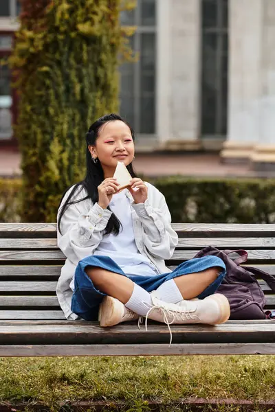 A stylish young woman relaxes on a park bench, savoring a snack, embodying youth culture at her university. — Stock Photo