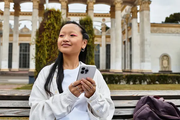 A fashionable Asian student relaxes on a bench, engaging with her smartphone amid a vibrant university atmosphere. — Stock Photo