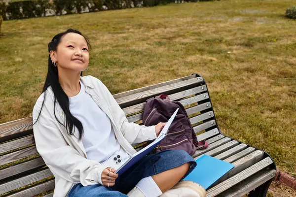 A stylish young Asian student sits on a bench, lost in thought while studying on her university campus. — Stock Photo