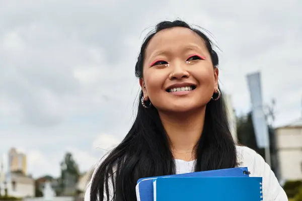 A young Asian girl with a joyful smile holds her notebooks while enjoying campus life. — Stock Photo