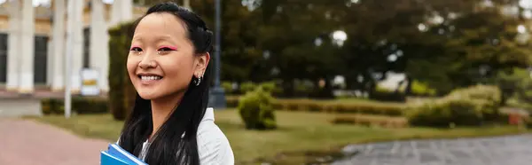 Um jovem estudante asiático está sorrindo brilhantemente enquanto mantém livros perto de sua universidade em um dia vibrante. — Fotografia de Stock
