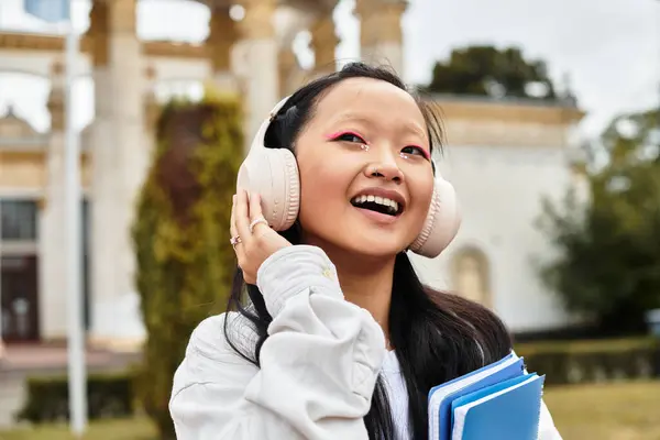 A young Asian woman in a stylish outfit smiles, listening to music on campus by her university in autumn. — Stock Photo