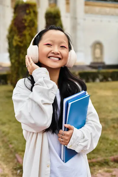 A cheerful student with headphones smiles brightly as she carries her notebooks near the university grounds. — Stock Photo