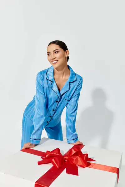 A young woman smiles delightfully while leaning on a large present adorned with a red bow. — Stock Photo