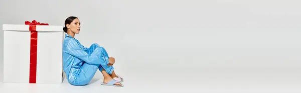 A young woman in stylish blue attire contemplates next to a large gift adorned with a red ribbon. — Photo de stock