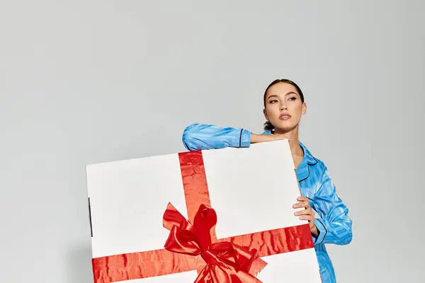 A joyful young woman dressed in blue showcases a big gift with a striking red ribbon indoors. — Stockfoto