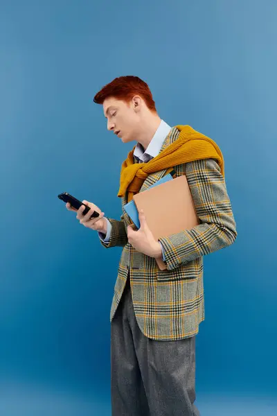 A young man in a checkered blazer and yellow sweater is focused on his phone while holding books. — Stock Photo