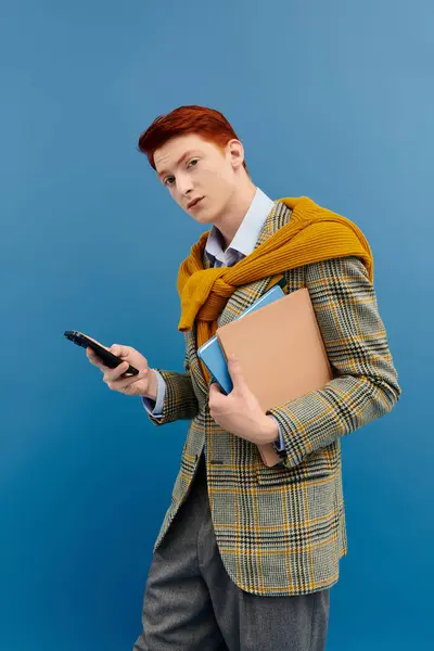 Um jovem estudante com cabelo ruivo mostra seu senso de moda em uma roupa de inverno enquanto segura livros e um telefone. — Fotografia de Stock