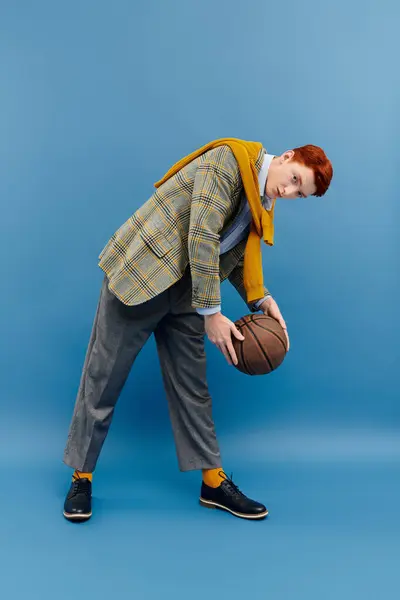 Un jeune homme habillé aux cheveux roux montre son sens de la mode tout en tenant un ballon de basket dans un studio. — Stock Photo