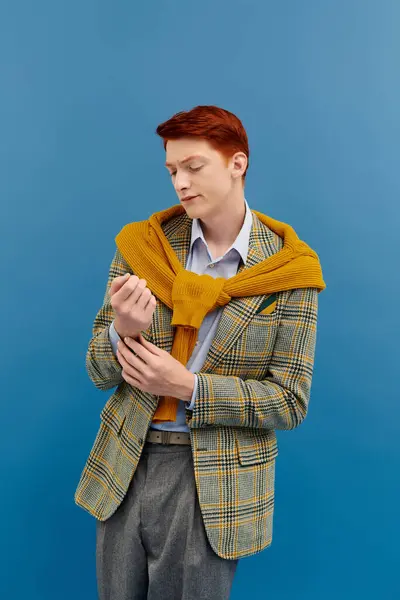 Un joven de moda con el pelo rojo vibrante posa con confianza en un traje de invierno elegante contra un entorno azul. - foto de stock