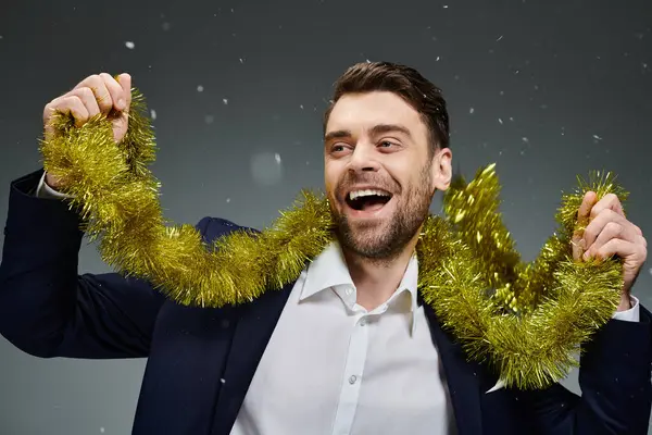 A cheerful young man wearing a suit joyfully showcases golden tinsel, embracing winter festivities. — Stock Photo