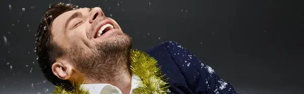 A handsome young man is filled with joy as snowflakes fall gently while he wears a festive garland. — Fotografia de Stock