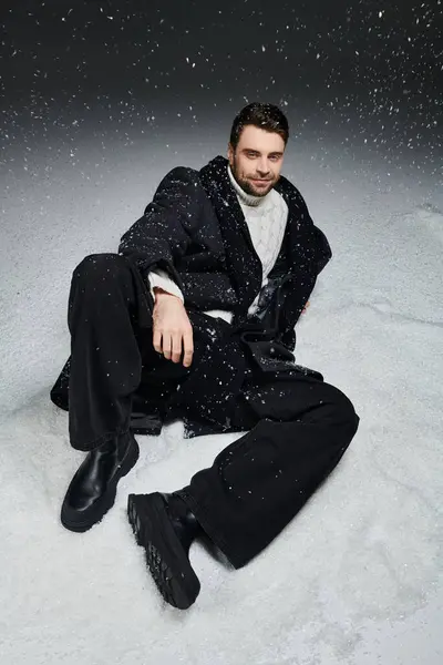 A handsome young man relaxes in a snowy setting, dressed in trendy winter attire while it snows. — Stock Photo