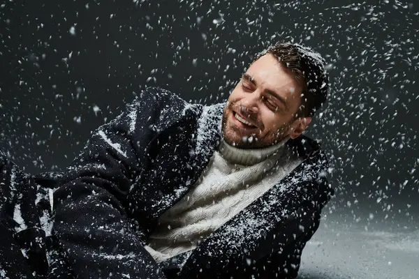 Un giovane uomo in un maglione spesso ride felicemente mentre i fiocchi di neve cadono delicatamente intorno a lui all'interno. — Foto stock