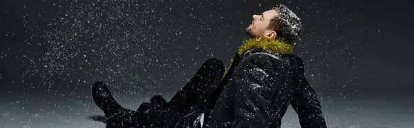 A stylish young man sits on the snowy ground with snowflakes falling around him. — Stock Photo