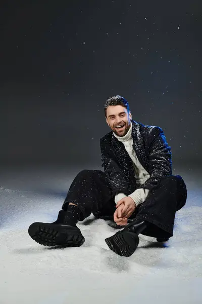 Este joven con un elegante atuendo de invierno se sienta juguetonamente en la nieve, exudando felicidad y encanto. - foto de stock