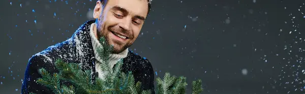 Snowflakes dance around a smiling young man gathering vibrant green branches on a winter day. — Stock Photo