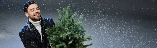 A cheerful young man embraces a Christmas tree while snowflakes swirl around him in winter joy. — Stock Photo