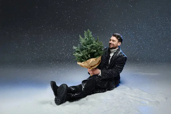 The young man smiles as he holds a festive tree while surrounded by falling snowflakes. — Photo de stock