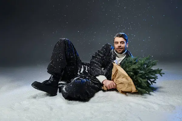 A young man lounges in the snow, holding a freshly cut Christmas tree, enjoying a winter moment. — Stock Photo