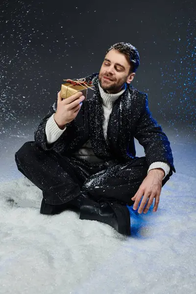 A young man sits on snow, smiling as he holds a wrapped gift in the winter atmosphere. — Stock Photo
