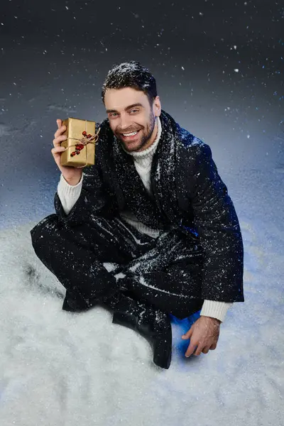 A cheerful young man dressed in winter attire sits in snow, holding a special gift with happiness. — Stock Photo