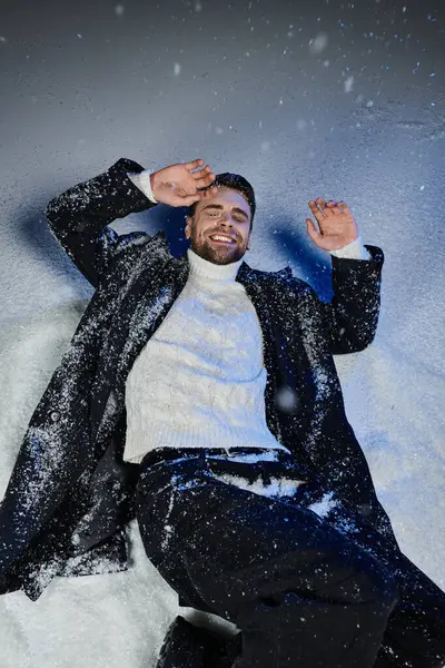 A handsome man smiles brightly while playfully lying in the snow, surrounded by winter scenery. — Stock Photo