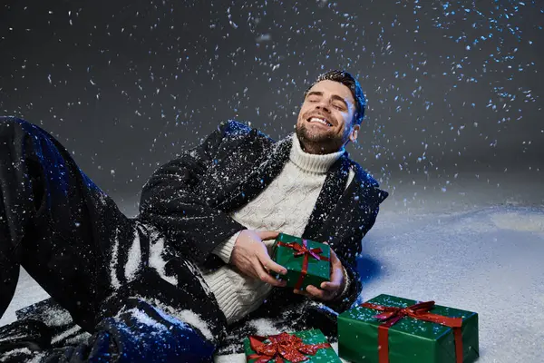 Cheerful young man sits on snow covered ground surrounded by presents while enjoying the moment. — Stock Photo