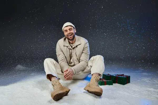 A young man sits in the snow, wearing warm clothing, laughing joyfully among colorful presents. — Stock Photo