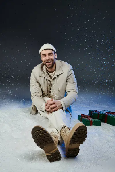 A cheerful young man sits in soft snow, surrounded by colorful gifts, radiating happiness. — Stock Photo