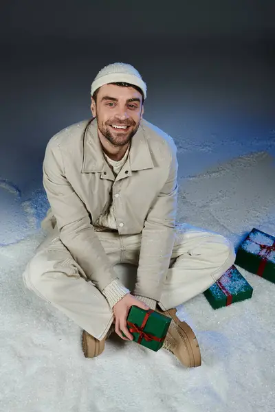 A smiling young man sits in the snow, joyfully holding a wrapped gift among decorations. — Stock Photo
