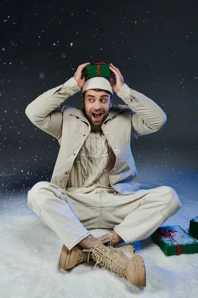 A cheerful young man sits on the ground, playfully balancing a gift while snow falls around him. — Stock Photo