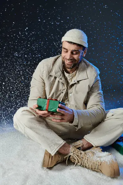Un joven alegre se sienta en la nieve, abriendo con entusiasmo un regalo maravillosamente envuelto. - foto de stock