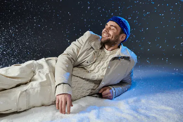 A charming young man relaxes in fresh snow, smiling joyfully amidst falling snowflakes. — Stock Photo
